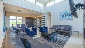 A spacious living room with a large black leather sofa, blue armchairs, and a grey sofa. There is a black coffee table with decorative items, a patterned rug, and a painting on the wall.