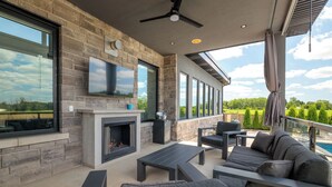 A modern outdoor living area with a stone fireplace, comfortable seating, and large windows offering a view of the landscape.