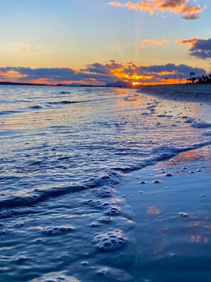 Et strandbillede med bølger, der forsigtigt slår mod kysten, en klar himmel og solen, der går ned i det fjerne.
