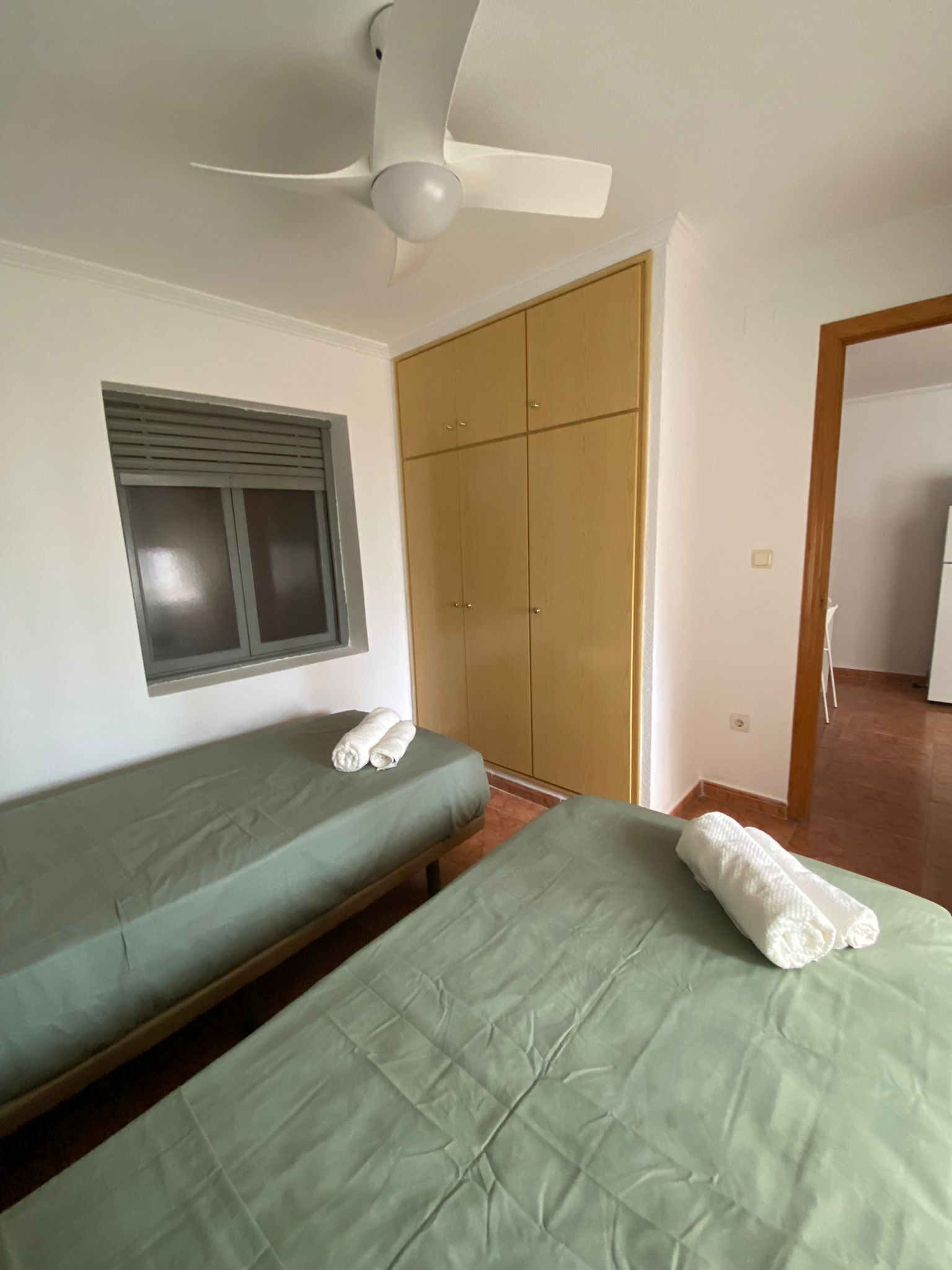 A room with two single beds, a wardrobe, a ceiling fan, a window with blinds, and a door leading to another room.