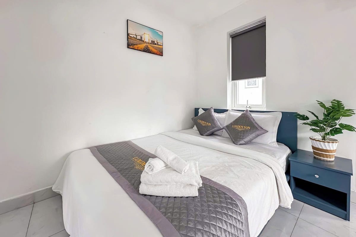Ein ordentlich bezogenes Bett mit einer grauen Decke, zwei Kissen mit dem Aufdruck 'Golden Sun', ein aufgerolltes weißes Handtuch und eine kleine Topfpflanze auf einem Nachttisch.