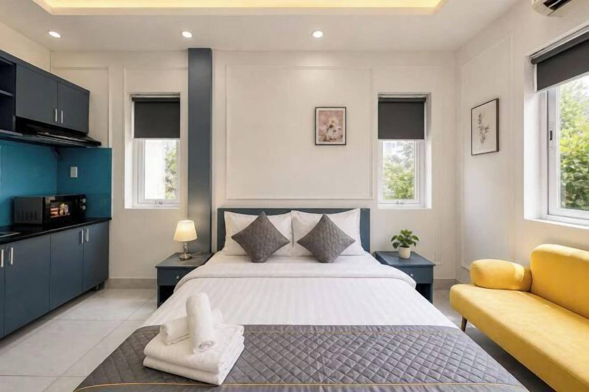 A modern hotel room with a bed, bedside tables, a sofa, a kitchen area, and a window with a view.