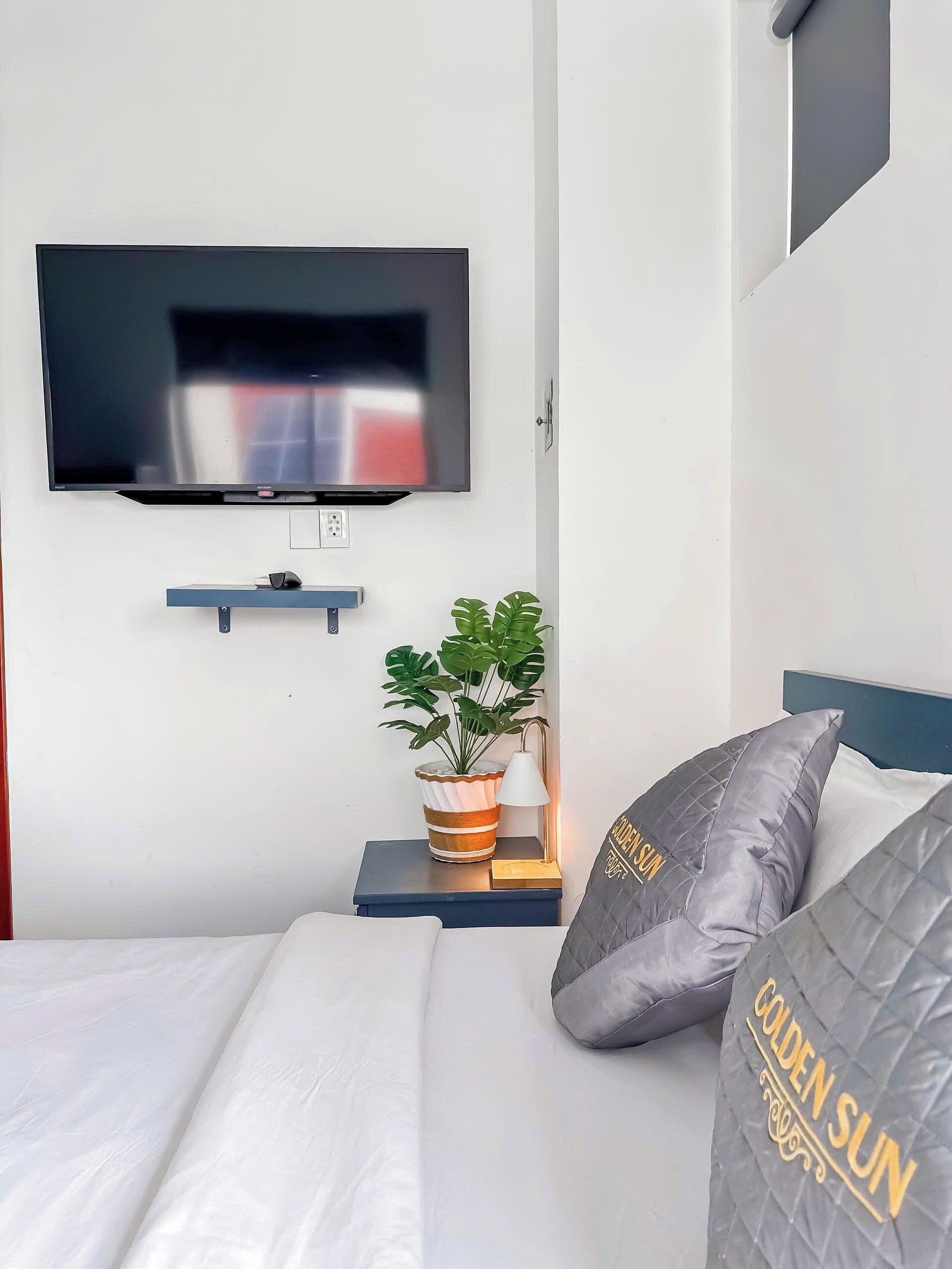 Moderni hotellihuone, jossa on seinälle asennettu taulutelevisio, yöpöytä, jossa on ruukussa oleva kasvi, ja sängyssä on tikattu tyyny, jossa on merkintä ”GOLDEN SUN”.