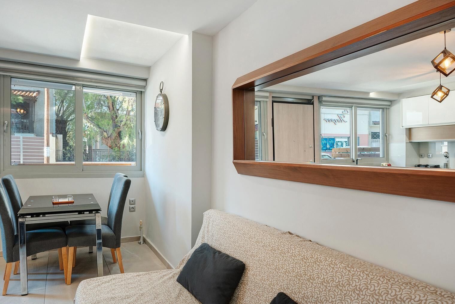 A modern living room with a sofa, a dining table with chairs, a large mirror, and a view of the outside through a window.