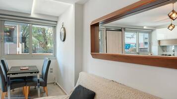 A modern living room with a sofa, a dining table with chairs, a large mirror, and a view of the outside through a window.