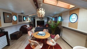 Een moderne woonkamer met een bank, een salontafel en een eethoek met stoelen. Er zijn meerdere ronde ramen in de wanden, een grote flatscreen televisie en een plafondlamp.