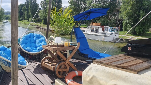 Een scheepsdek met een blauwe fauteuil met kussens, een houten tafel en een blauwe parasol.