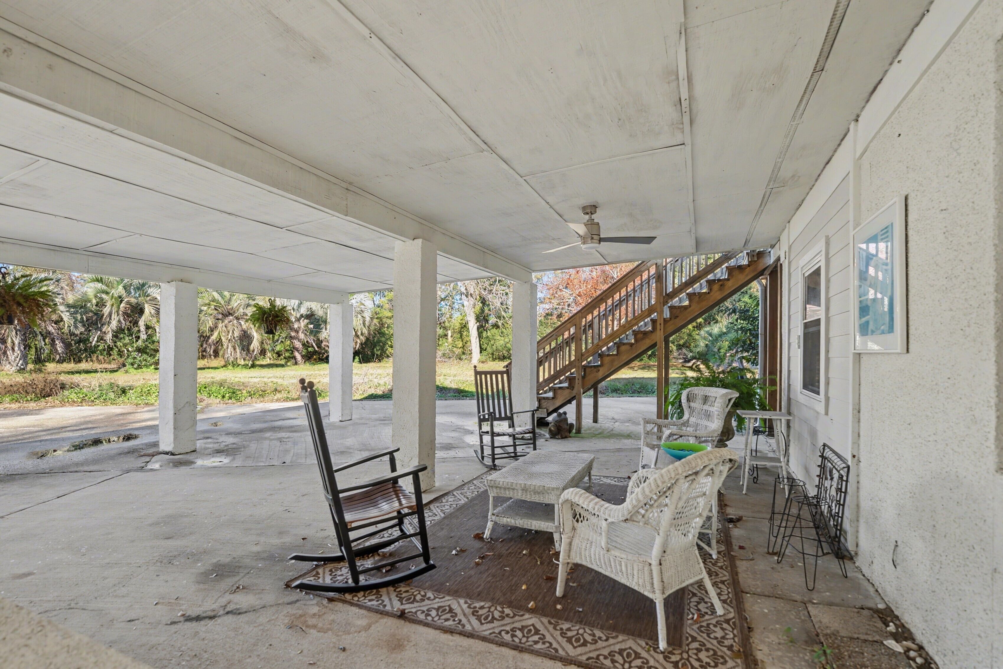 An outdoor covered patio with white wicker furniture, a concrete floor, and a wooden staircase leading to a building.