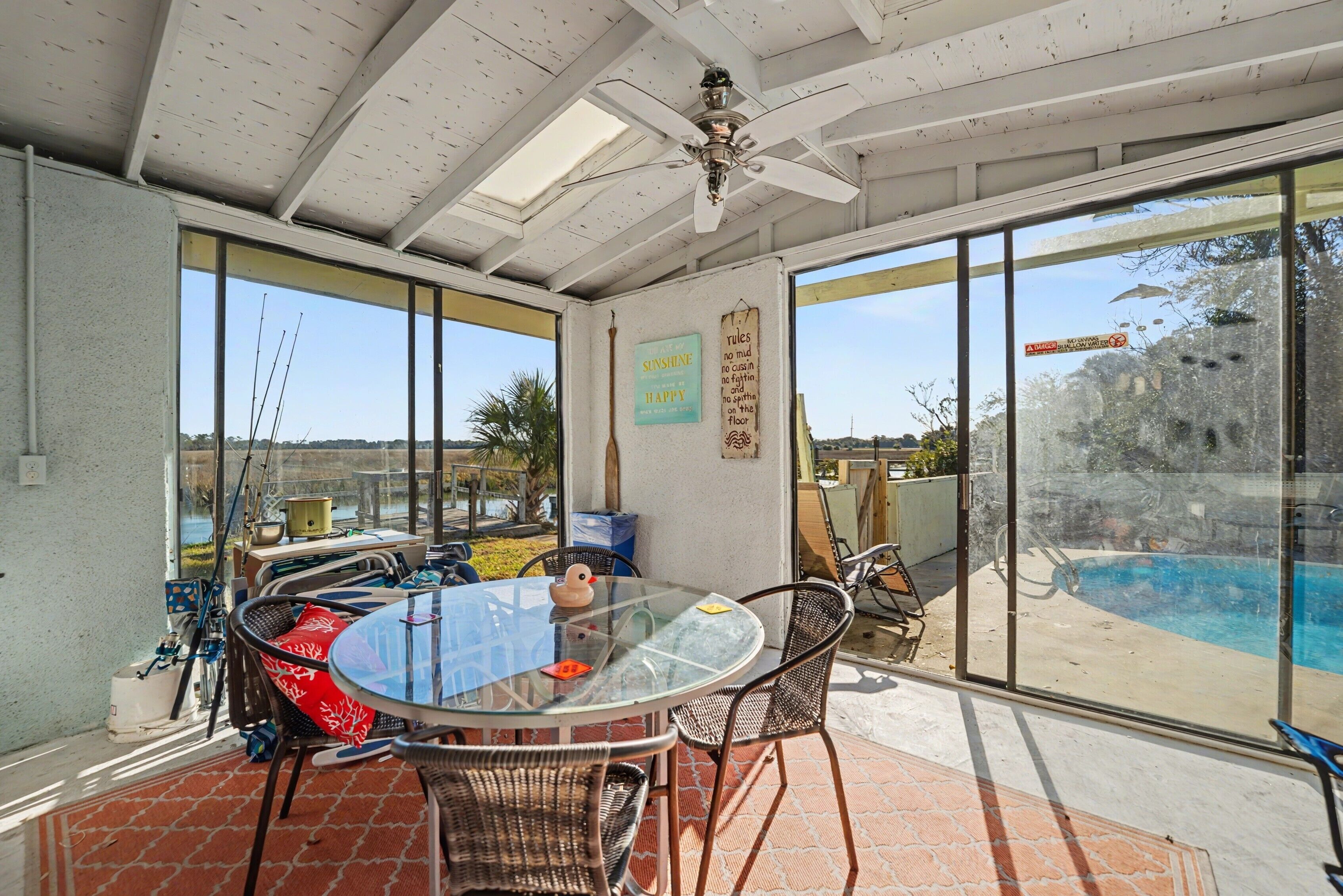 A sunlit patio with a glass table and chairs, a ceiling fan, and a view of a swimming pool.