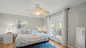Ein Schlafzimmer mit Bett, Nachttischen, Deckenventilator und einem großen Fenster mit Vorhängen.