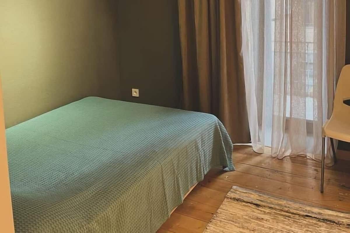 Ein Zimmer mit einem Einzelbett, einem Sessel und einem Fenster mit Vorhängen.