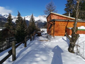 Une cabane en bois, avec un sentier enneigé menant à celle-ci, entourée d’arbres et de montagnes.