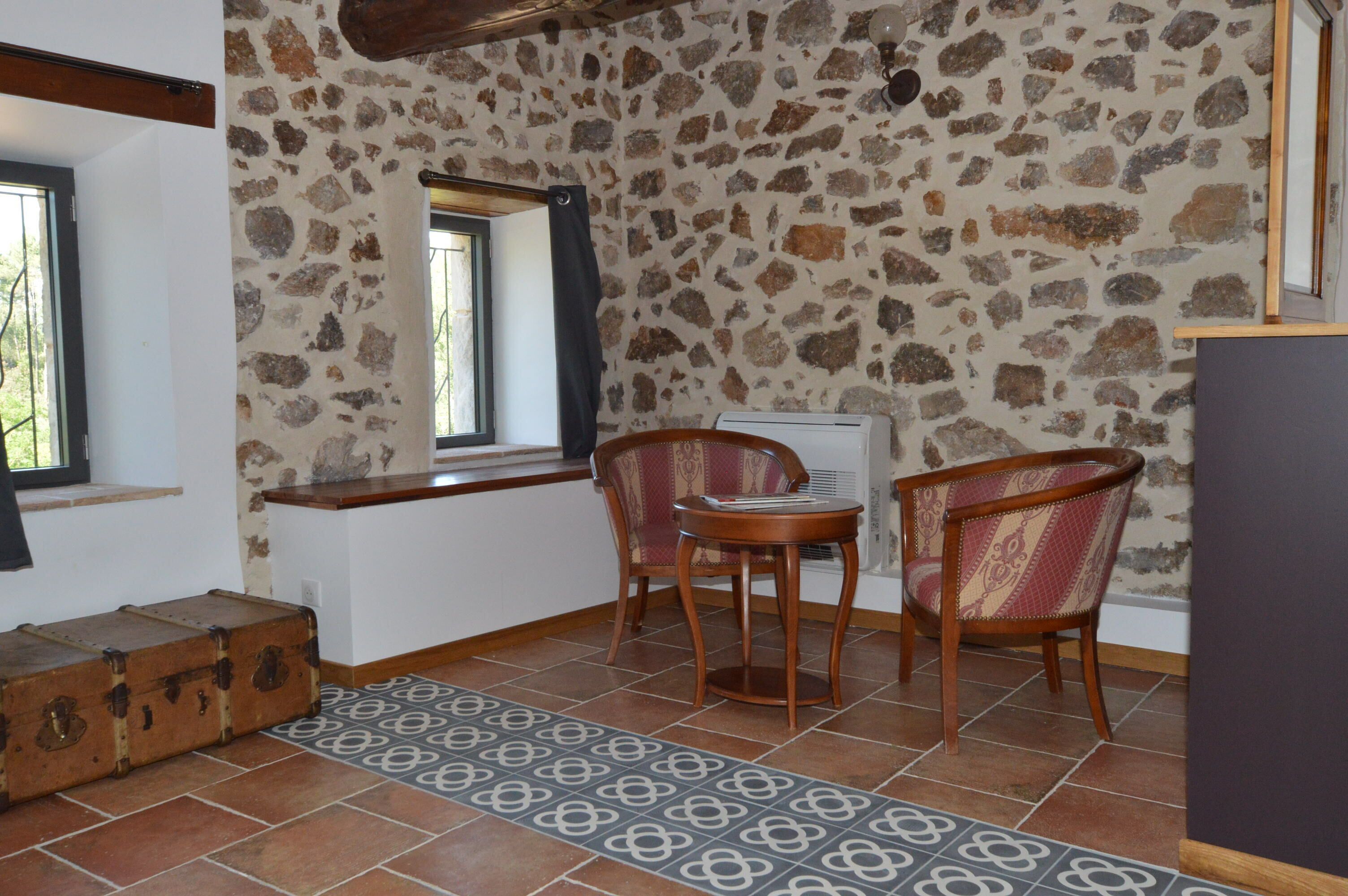 Una habitación con pared de piedra, una ventana, un banco de madera, dos sillas, una alfombra estampada y una maleta.