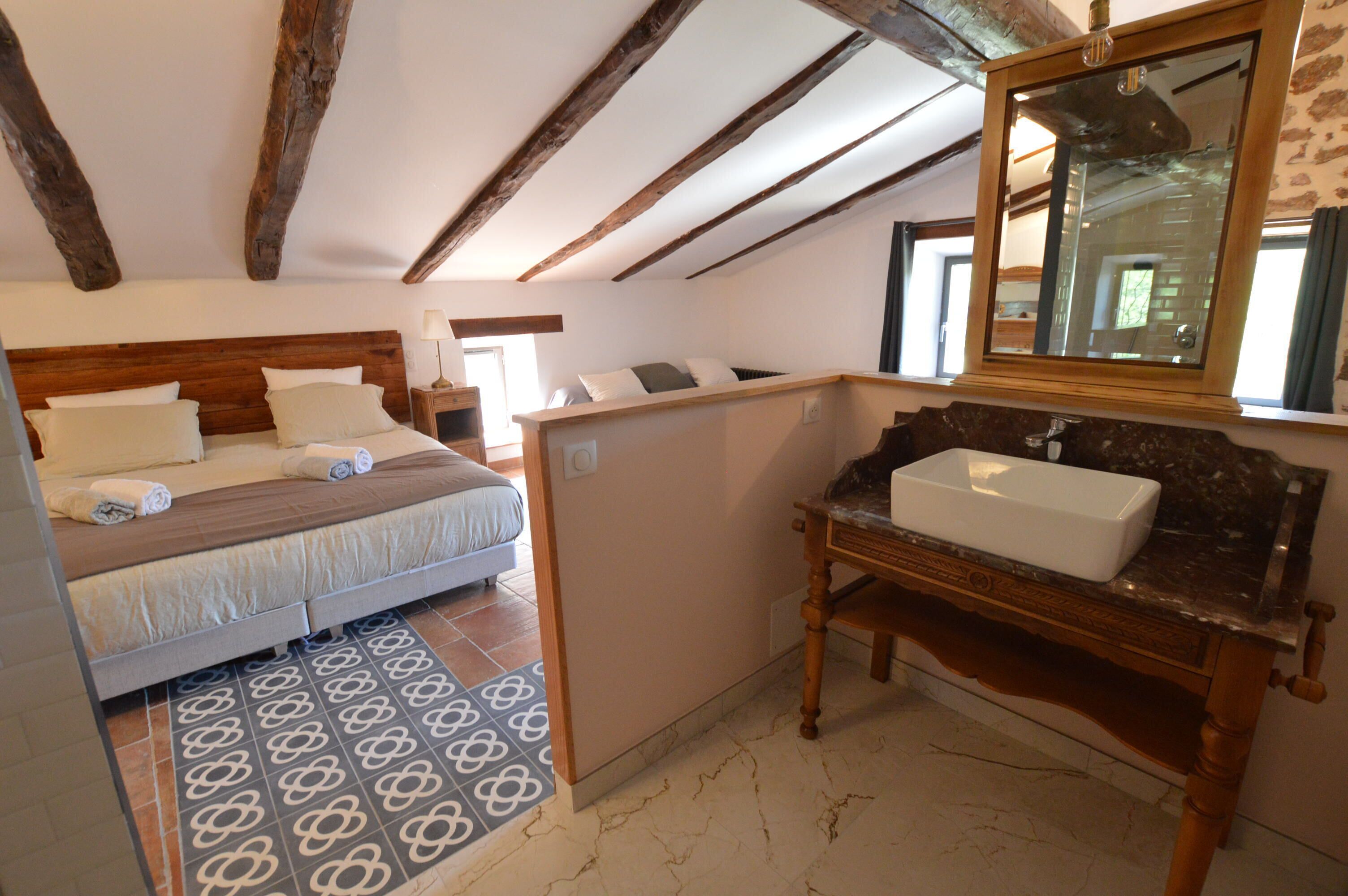 Een slaapkamer met een houten hoofdbord, een bed met kussens, een nachtkastje, een spiegel, en een badkamer met een wastafel en marmeren vloer.