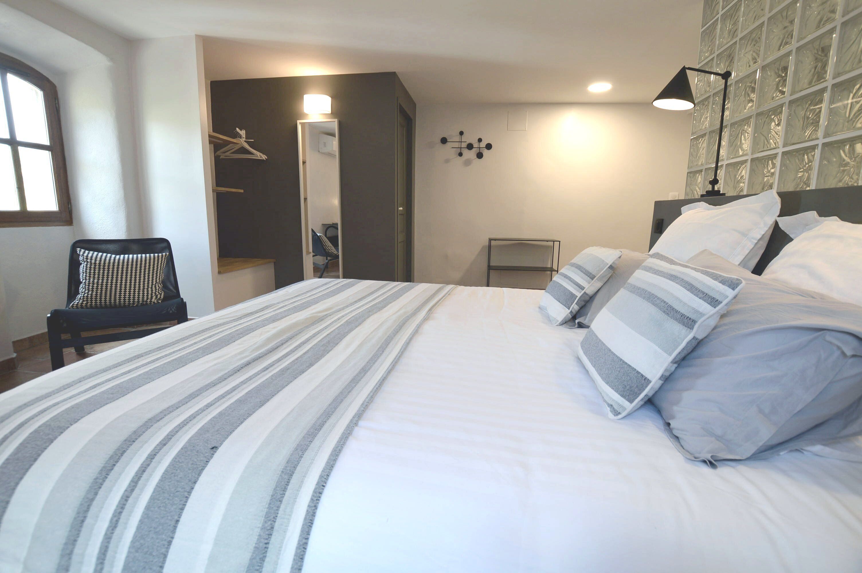 A neatly made bed with striped bedding, a chair with a checkered cushion, and a mirror with a light above it.