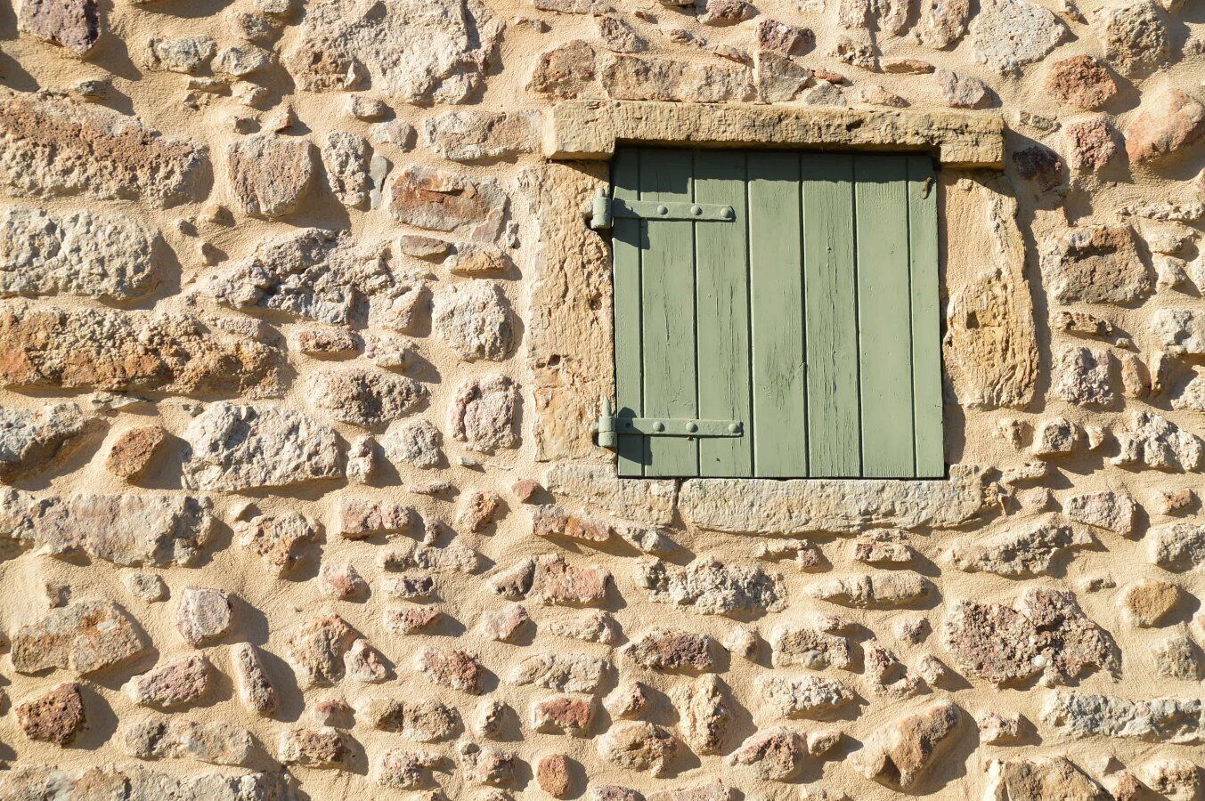 Un muro de piedra con un pequeño ventanal y un persiano verde.