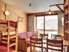 A cozy cabin interior with a bunk bed, a sofa, a dining table, and a large window offering a view of the outdoors. - 200m from resort center, 12nd floor, view ski slopes, swimming pool, balcony, tv, ski locker, 22m² (Le Corbier)