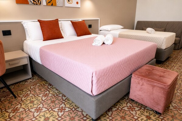 Habitación de hotel con cama, mesita de noche, silla, pequeña mesa, otomana rosa y baldosas de suelo estampadas.