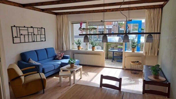 Un salón acogedor con un sofá azul, una silla de madera, una mesita y una mesa de comedor. Hay plantas en macetas y se ve la calle desde el interior.