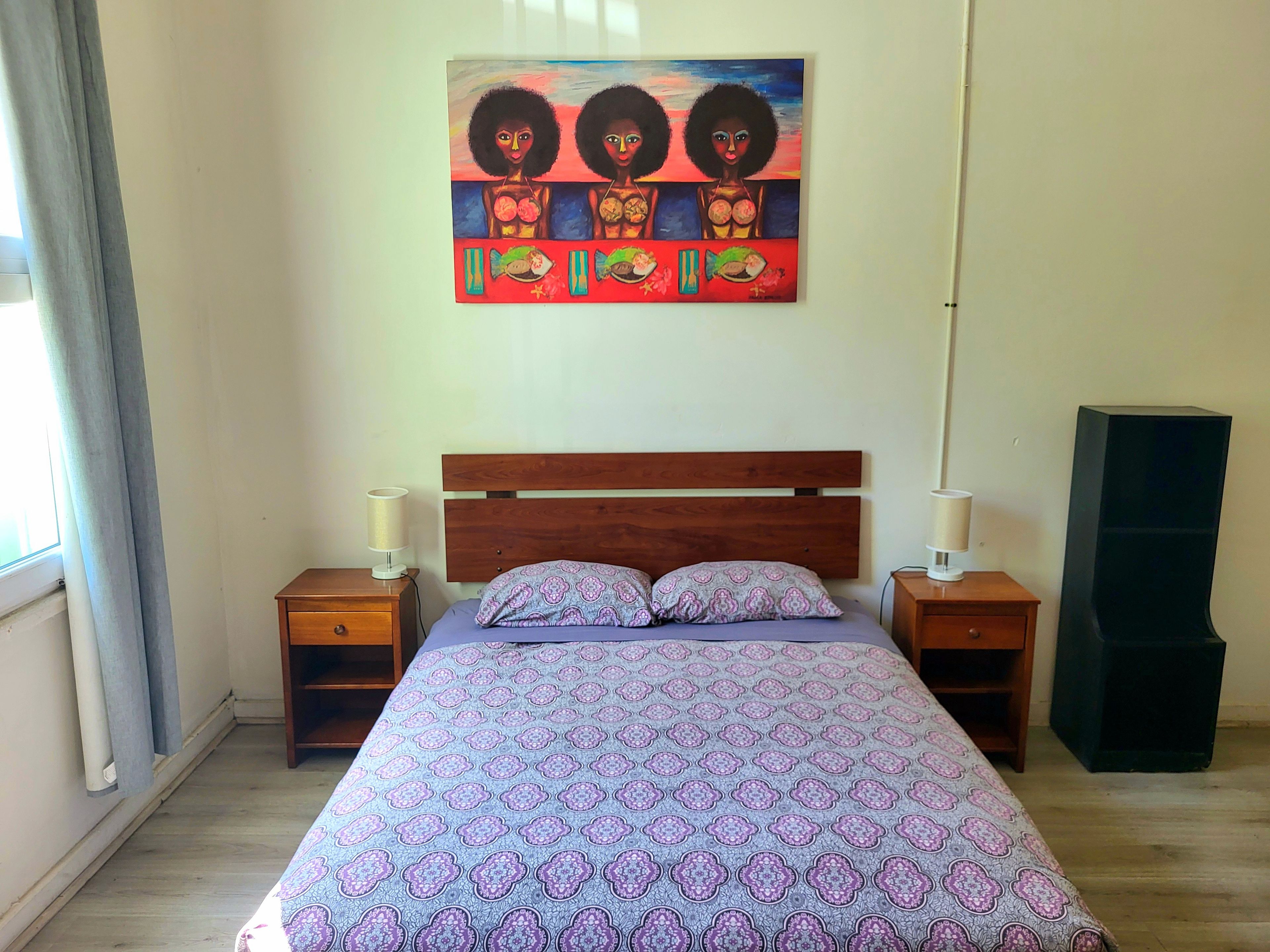 A bedroom with a wooden bed, two bedside tables, a wall-mounted painting, and a tall black speaker.
