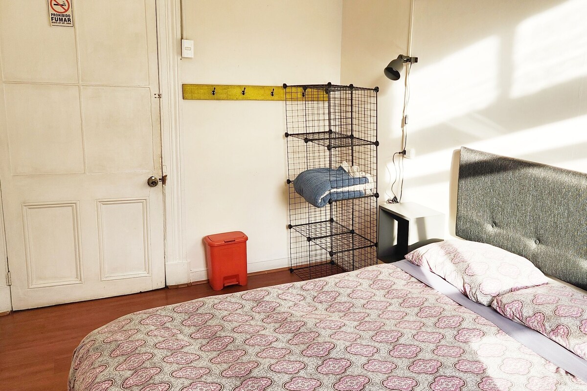 A bedroom with a bed, a wardrobe, a small stool, a shelf with clothes, and a wall-mounted lamp.