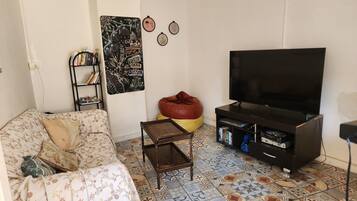 Uma sala de estar com sofá, mesa de centro, um puff, televisão, estante de livros e um objeto decorativo na parede.