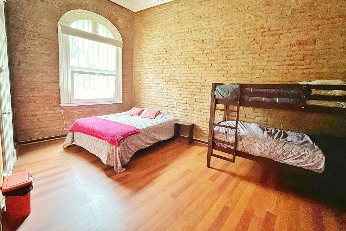 A room with a bunk bed, a single bed with a pink blanket, a window with white frames, and a brick wall.