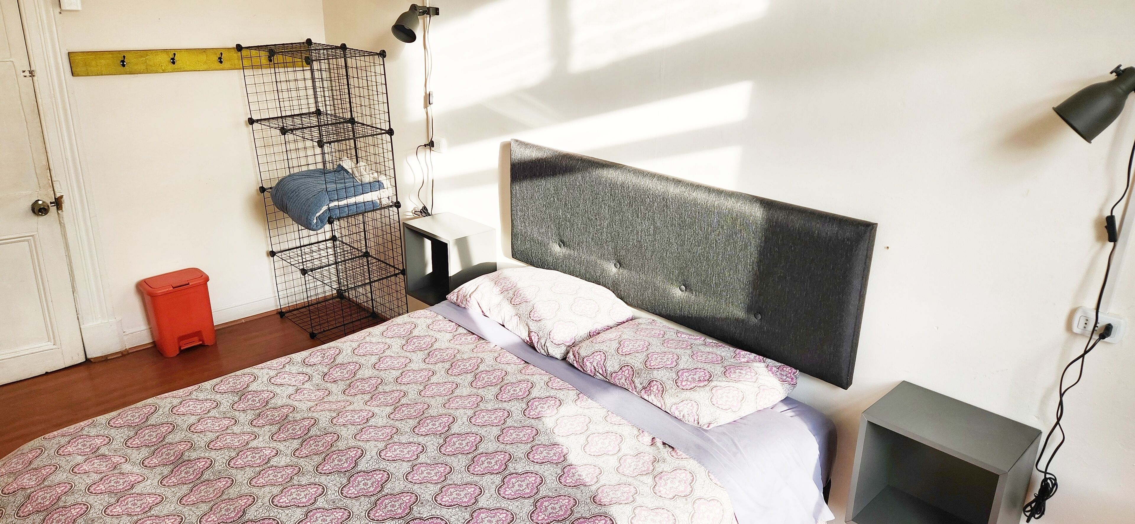 Ein Schlafzimmer mit Bett, einem Kopfteil, einem an der Wand befestigten Regal, einer roten Mülltonne und einem Schreibtisch mit Lampe.