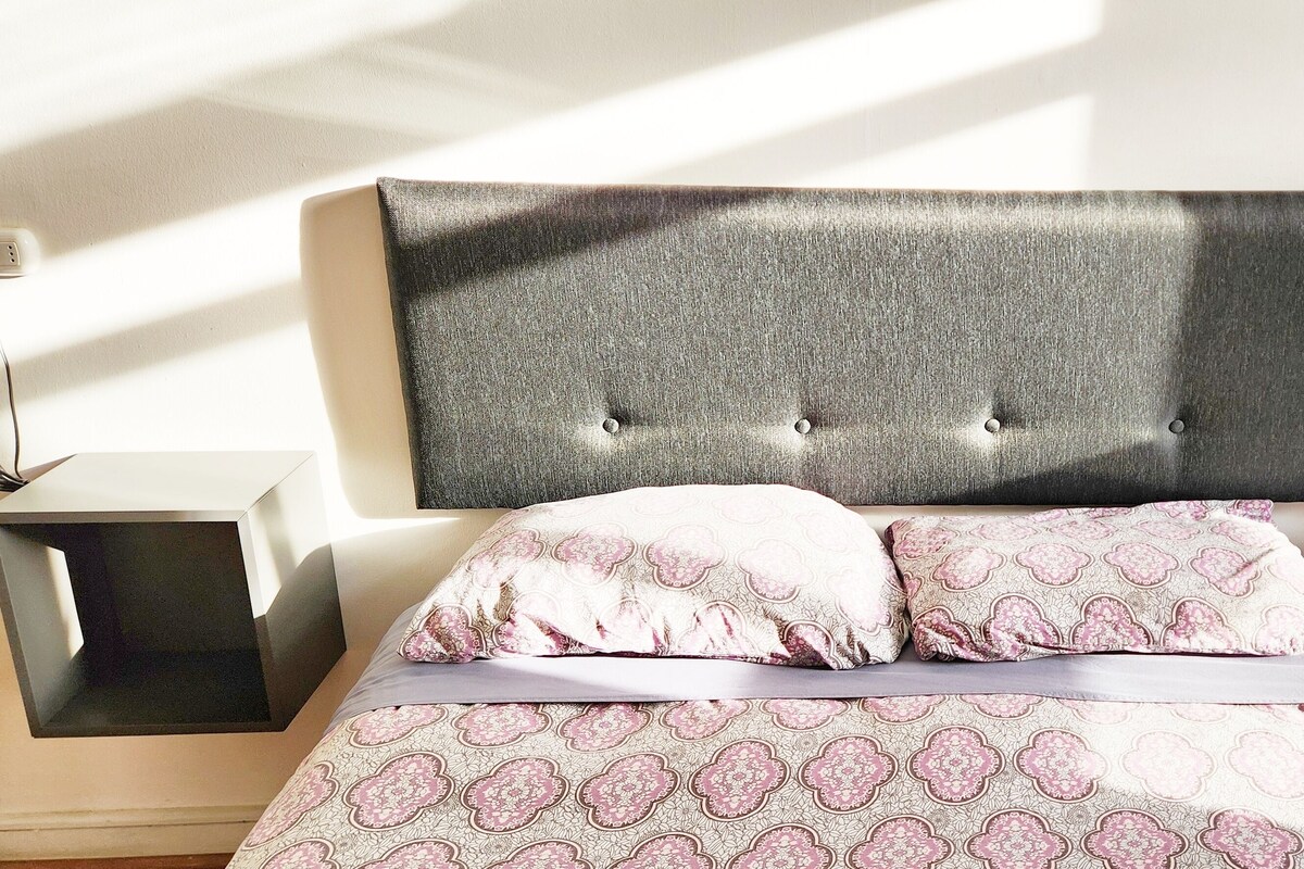 A neatly made bed with a patterned bedspread and pillows, flanked by two bedside tables with black square bases.
