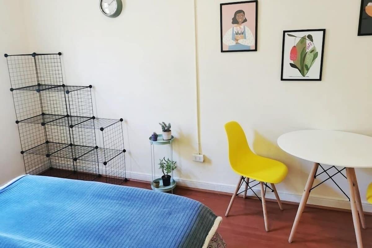 A room with a bed, a yellow chair, a white table, a clock, and two framed pictures on the wall.