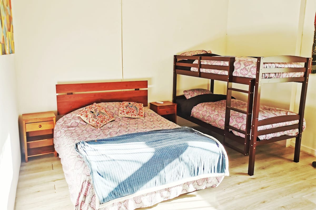 A bunk bed with a wooden headboard and a lower bed with patterned bedding. There is a wooden nightstand with a drawer and a small table beside the lower bed.