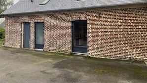 A brick building with two windows and a door, a sloped roof, and a paved ground.