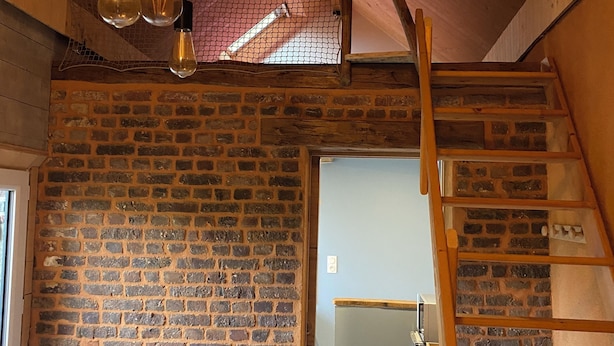A rustic interior with exposed wooden beams, a brick wall, and a loft area with a wooden ladder.
