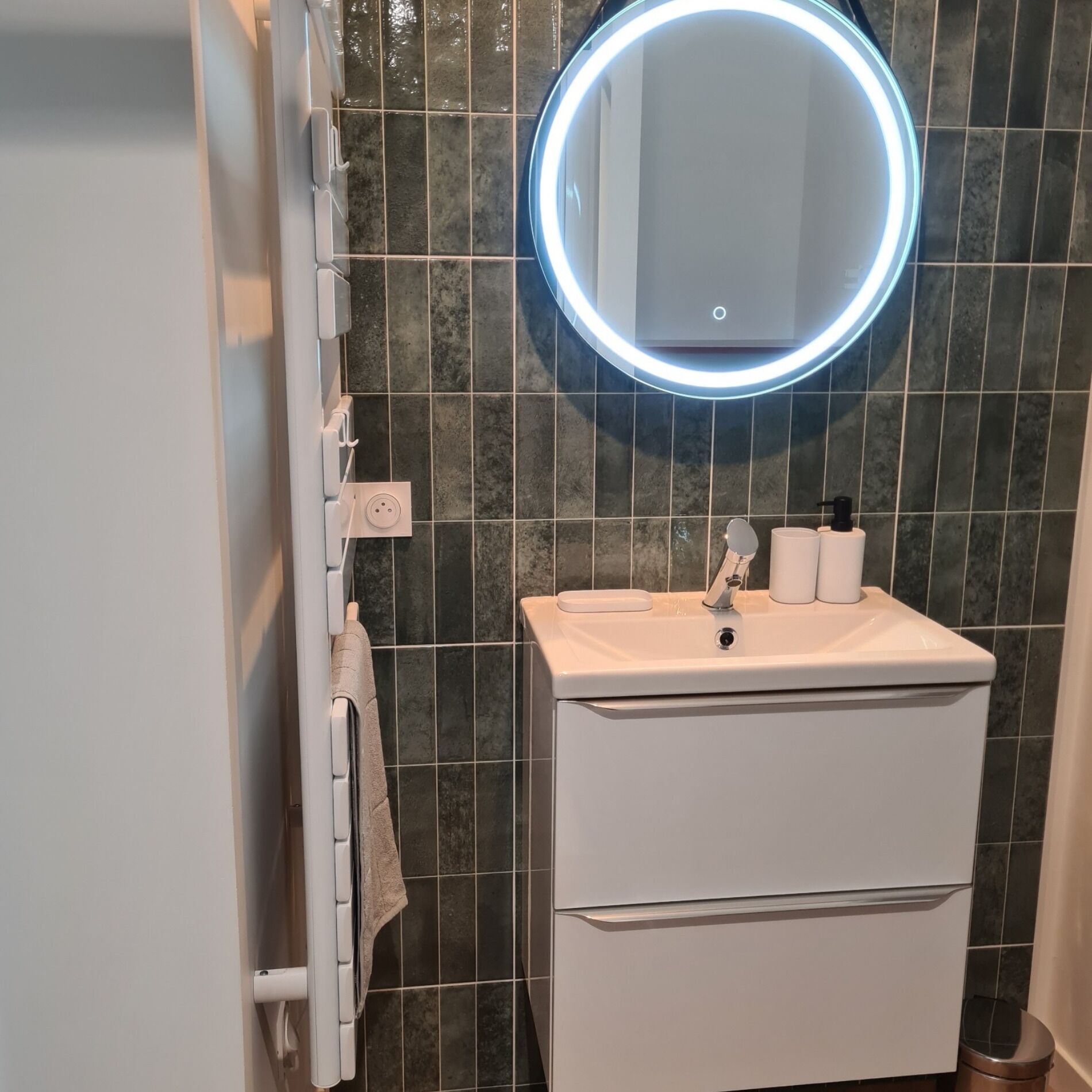 A bathroom with a white sink, a mirror with a circular light, and a towel hanging on a rack.