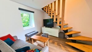 A modern living room with a grey sofa, colorful cushions, a wooden coffee table, a TV on a white cabinet, and a wooden staircase. - La Maison du Vieux Bourg (Perrier)