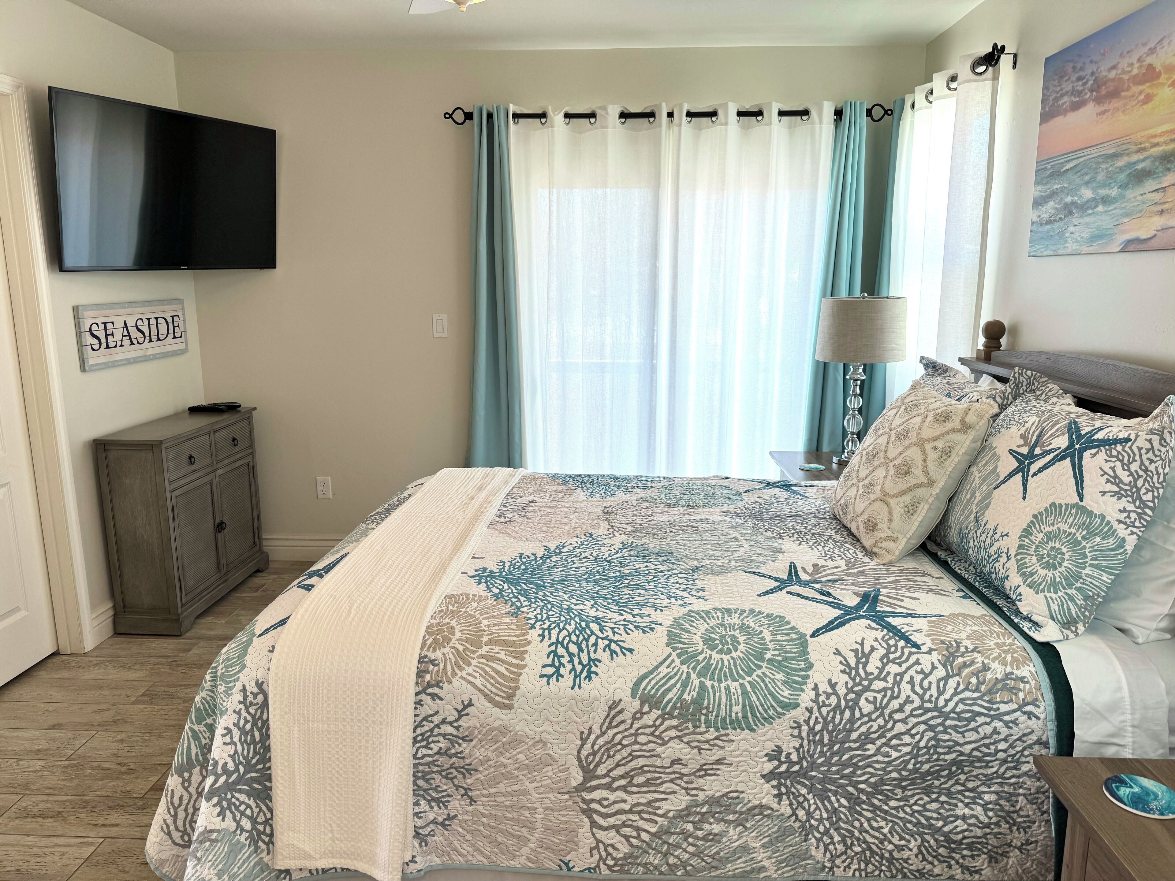 A bedroom with a bed, a TV mounted on the wall, a nightstand with a lamp, and a sign that reads "SEASIDE".