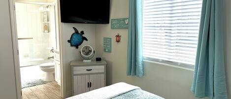 A bedroom with a bed, a TV mounted on the wall, a window with curtains, a small cabinet, and a wall decoration.
