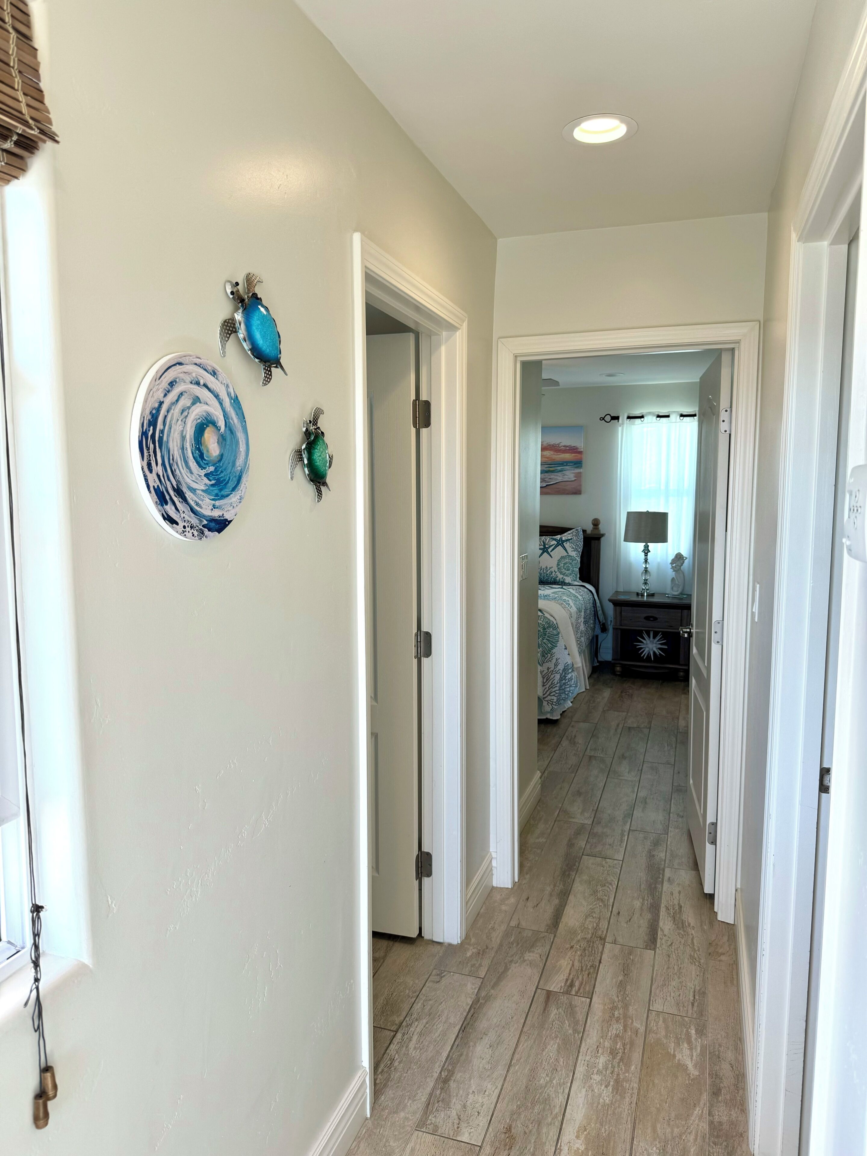 A hallway with a wooden floor, white walls, and a door leading to a bedroom. There are decorative wall art pieces and a window with blinds.
