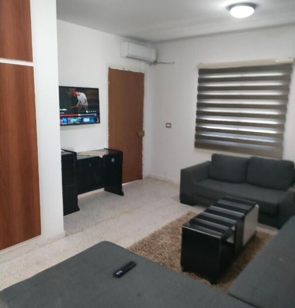 A living room with a grey sofa, a TV mounted on the wall, a wooden door, and a window with blinds.