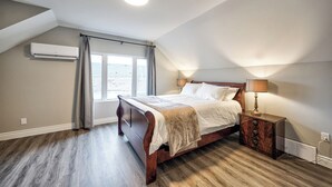 Une chambre avec un plafond en pente, un lit doté d’une tête de lit en bois, une table de chevet avec une lampe et une fenêtre avec des rideaux.
