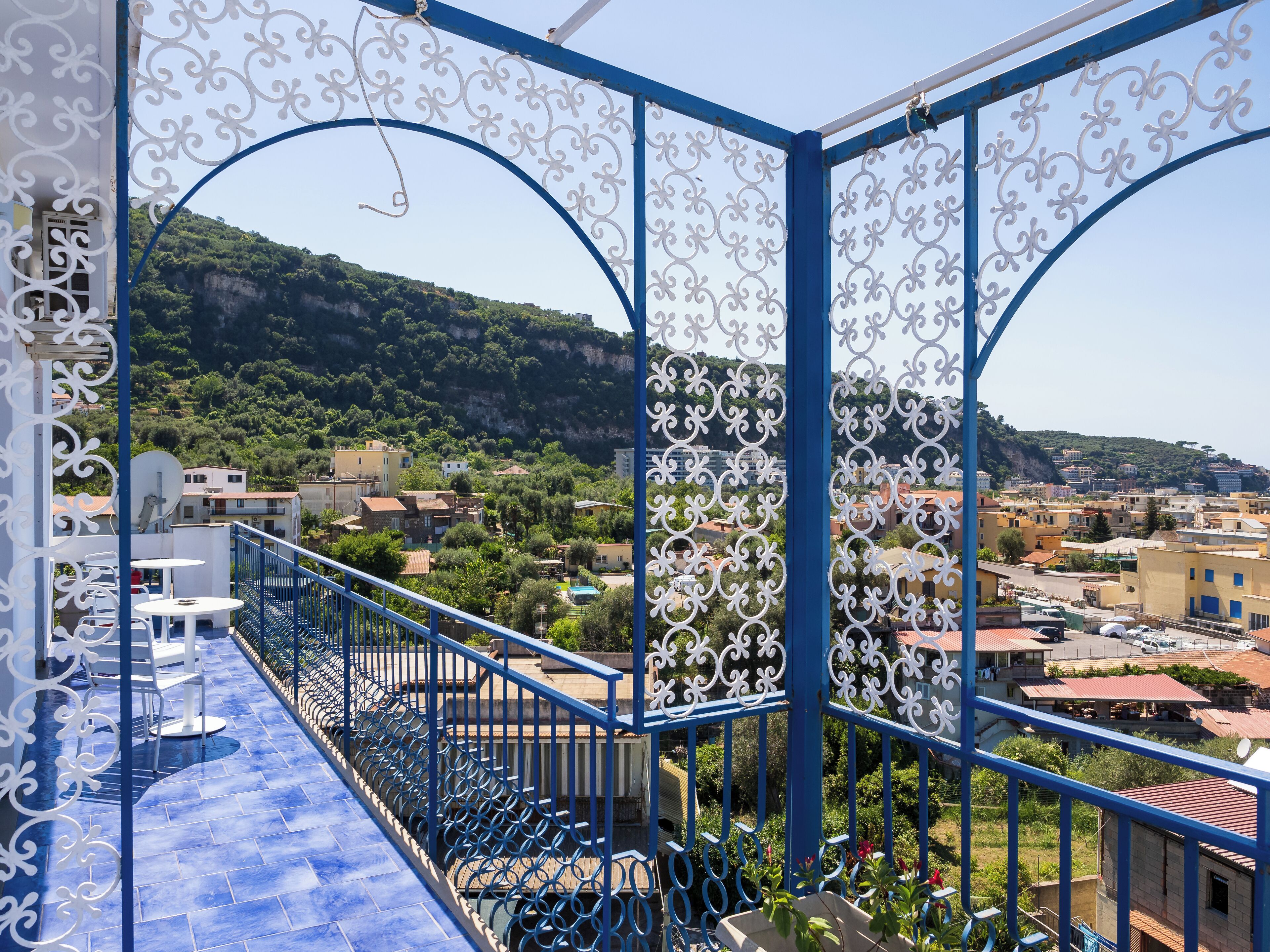 A balcony with a blue railing and white decorative elements, overlooking a hillside with buildings and greenery.