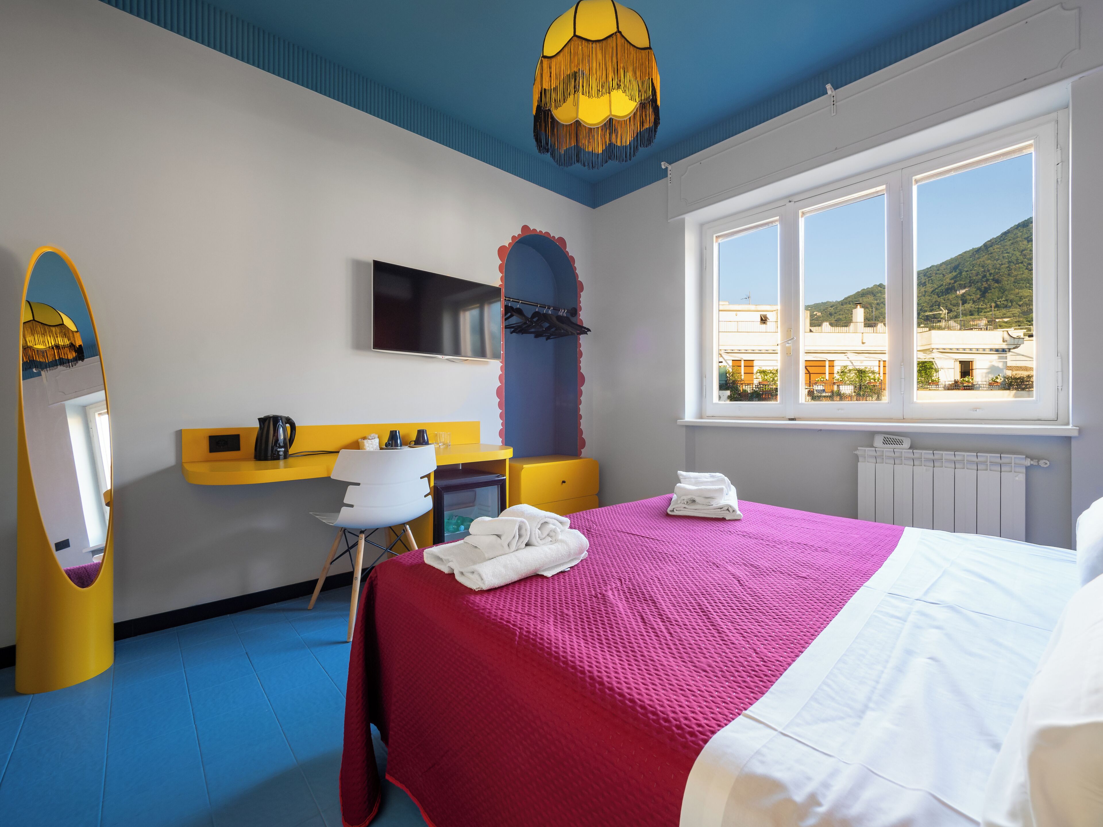 A hotel room with a bed, a desk, a chair, a mirror, a TV, and a window with a view of buildings and hills.