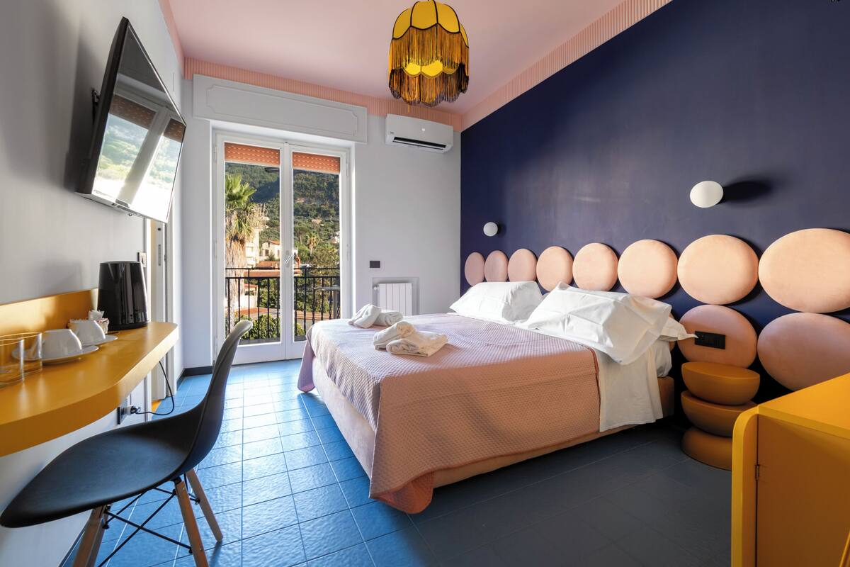 Een hotelkamer met een bed, een bureau, een stoel, een televisie en een balkon met uitzicht op de heuvels.