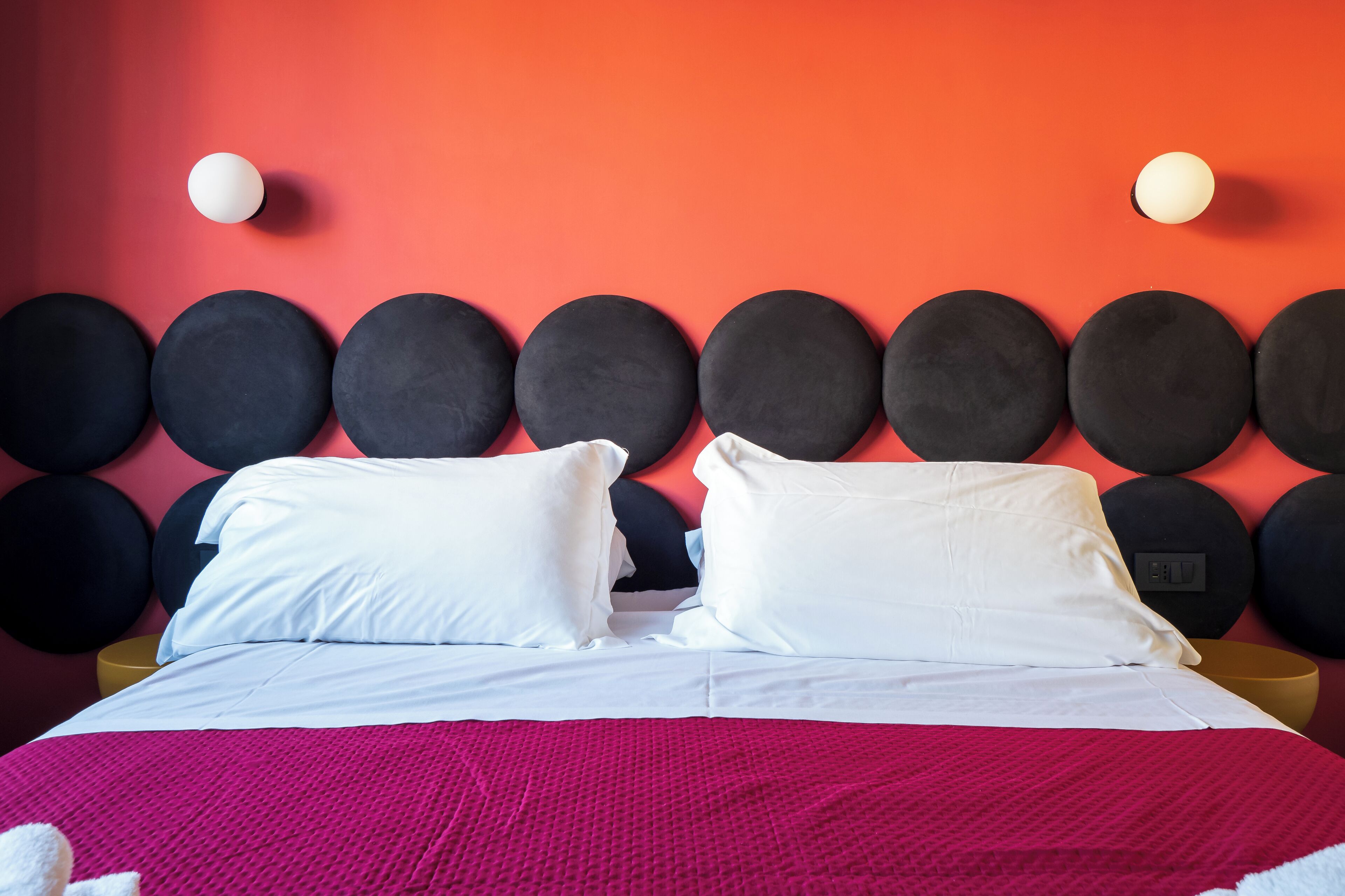 A bed with white linens and a pink blanket, a headboard with circular black and white accents, and a yellow side table.
