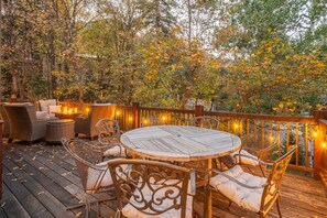 A wooden deck with a round table and wicker chairs, surrounded by trees and string lights.
