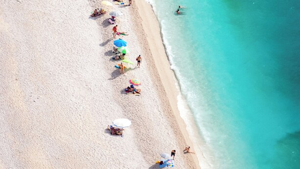 An aerial view of a beach with people swimming and sunbathing, and several colorful umbrellas.
