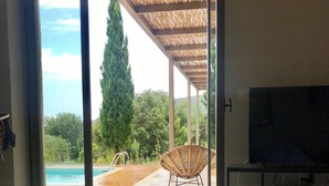 A modern living room with a large sliding glass door leading to a pool area, a wicker chair, a round ottoman, a television, and a coffee table with a tray.