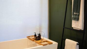 A modern bathroom with a freestanding bathtub, a wooden tray with toiletries, and a towel rack against a green wall.