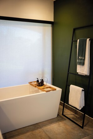 A modern bathroom with a freestanding bathtub, a wooden tray with toiletries, and a towel rack against a green wall.