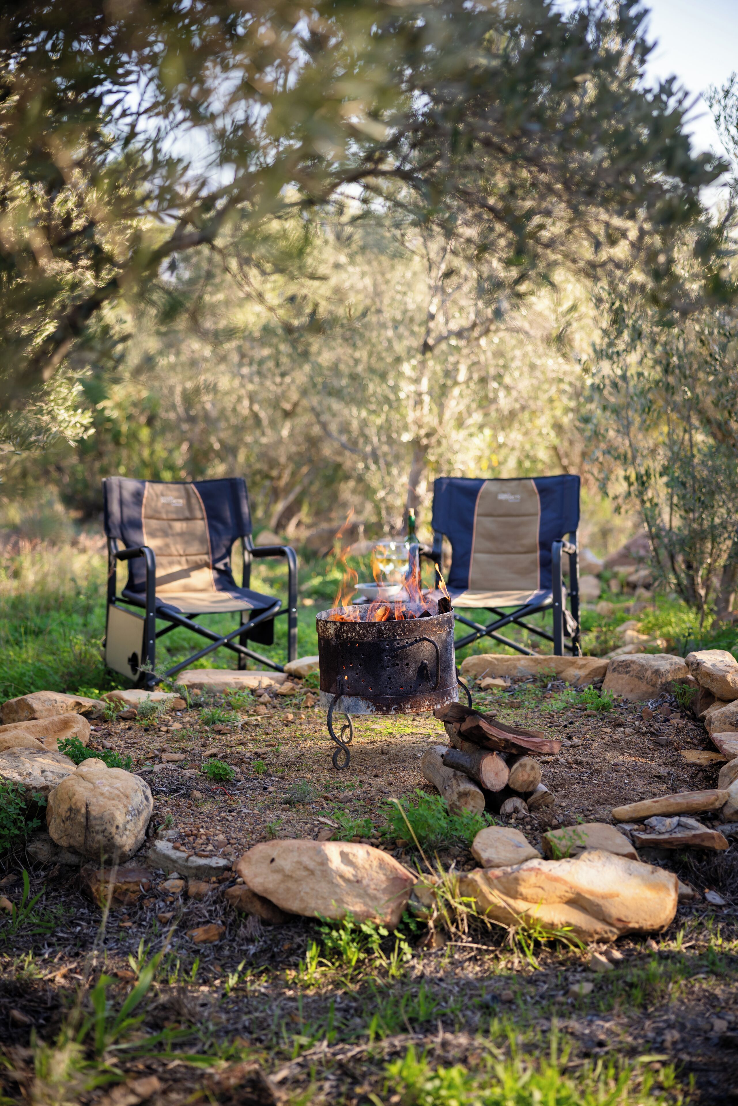 Two camping chairs with a campfire between them, surrounded by rocks and greenery.